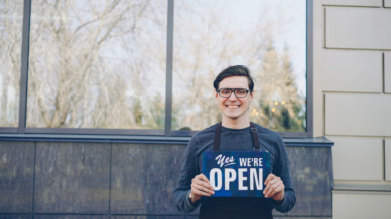 Smiling small business owner holding a "Yes, we're open" sign in front of their shop, the kind of local Chatham-Kent business that benefits from local SEO